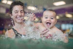 Mother holding a smiling baby boy in front of her as he splashes in the pool