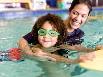 Boy with green goggles in a pool holding a kickboard learning to kick with swim instructor