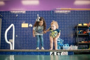Two girls holding hands in front of a swimming pool looking down at the water at Emler's Fort Worth Swim School location