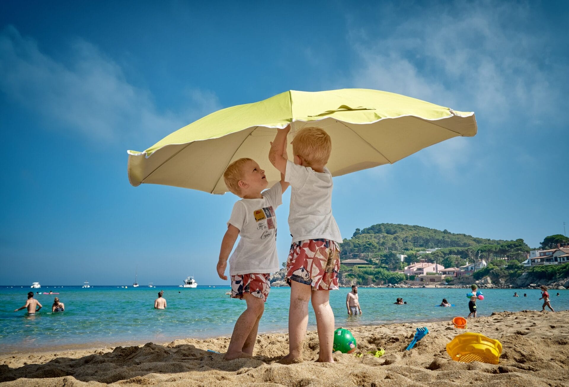 Two boys at the beach putting up an umbrella while families swim in the background
