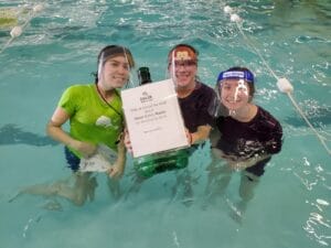 Three swim instructors in the pool with face shields holding a charity donation bucket