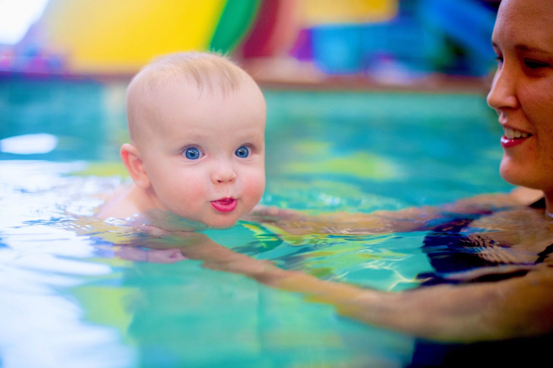 Blue-eyed baby being held at arms length learning to swim with instructor