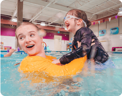 Instructor in yellow shirt holding a laughing girl as she teaches her to swim in the pool